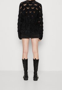 Pull noir et duveteux avec un motif en tricot ouvert, associé à une jupe en denim noir et des bottes en cuir noires jusqu'aux genoux, vu de derrière.