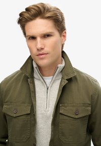 Chaqueta verde oliva con dos bolsillos frontales, cierre de botones y un suéter gris de cremallera debajo. Textura suave y ajuste a medida.