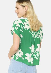 Camicia a maniche corte in tessuto verde con grandi motivi floreali bianchi, vestibilità comoda e dettaglio plissettato sul retro.