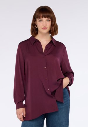 Mujer con cabello castaño hasta los hombros, usando aretes de plata en forma de aro, una camisa suelta burdeos de botones y jeans azules, con la mano en el bolsillo.