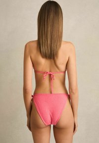 Růžové bikiny s texturovanými vzory listů, s tenkými ramínky uvázanými na zádech a po stranách, které mají bezešvý design a minimální zakrytí.