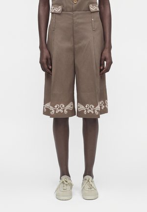 Barna, bőszárú rövidnadrág fehér hímzett részletekkel a zsebeknél és az alján, fehér tornacipővel viselve sötét bőrű lábakon.