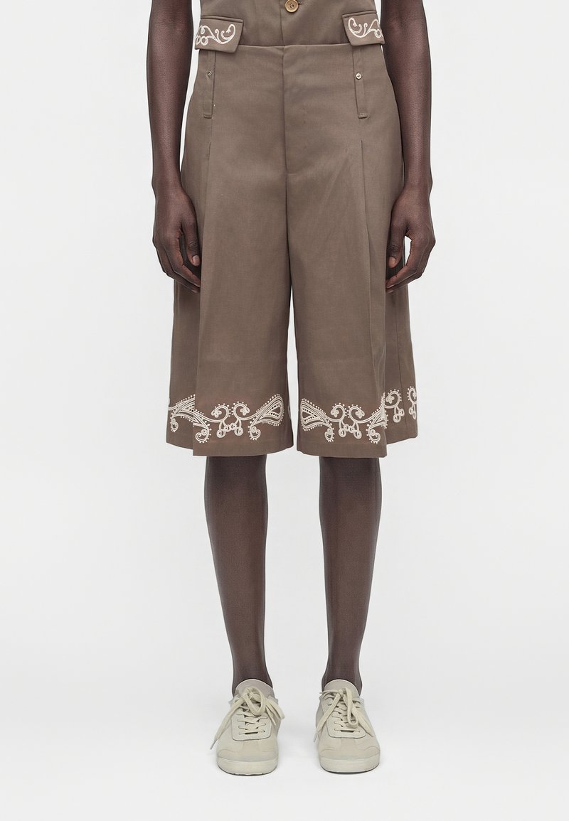 Καφέ σορτς με φαρδιά πόδια και λευκές κεντητές λεπτομέρειες στις τσέπες και τα τελειώματα, φορεμένα με λευκά αθλητικά παπούτσια σε σκούρα πόδια.