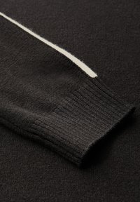 Zbliżenie czarnego swetra z długim rękawem, z białym paskiem przebiegającym wzdłuż rękawa oraz detalami ściągacza w formie prążków.