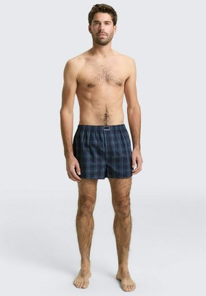 Erwachsener Mann steht barfuß, trägt dunkelblaue karierte Boxershorts und blickt nach vorn vor einem schlichten hellen Hintergrund.