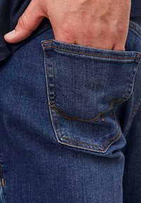 Sininen farkkuhousut, joissa on näkyvä etutasku, kontrastiväristä oranssia tikkausta, hieman kulutettu pinta ja rento istuvuus.