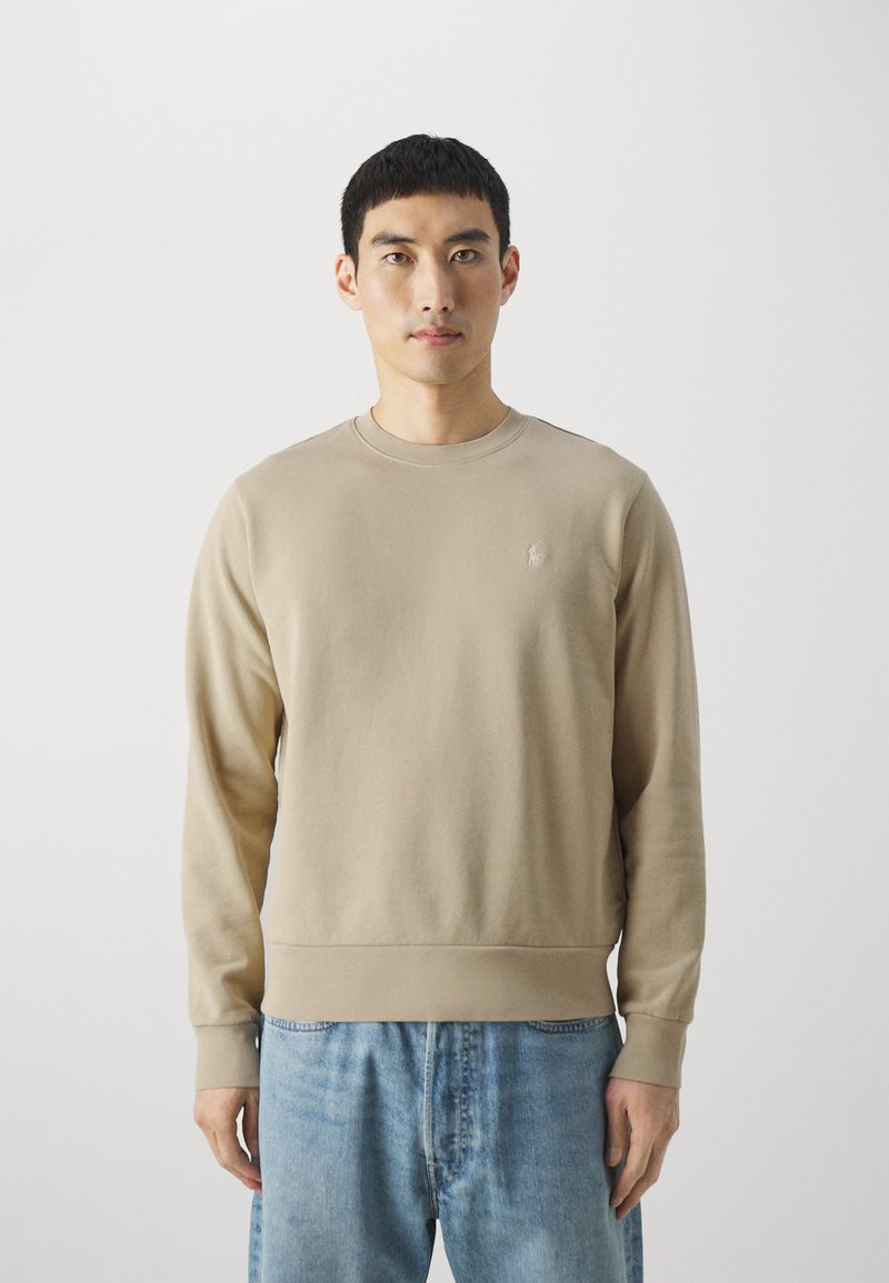 Beige crew-neck svetari valmistatud pehmest kangast, millel on pikad varrukad ja soonikuga äär. Rinnal on väike logo.