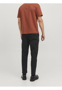 T-shirt di cotone color ruggine a maniche corte, abbinato a pantaloni neri a sigaretta. I pantaloni hanno una texture liscia e una leggera lucentezza. Scarpe nere completano il look.