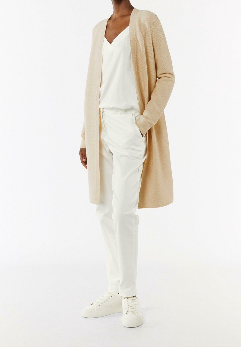 Beżowy długi kardigan na białym topie z dekoltem w serek, w zestawie z białymi spodniami i białymi tenisówkami. Gładka tekstura, luźny krój, minimalistyczny design.