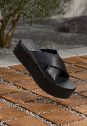 Black leather platform sandals with crossed straps and an elevated sole. Features a smooth finish and a subtle logo on the insole.