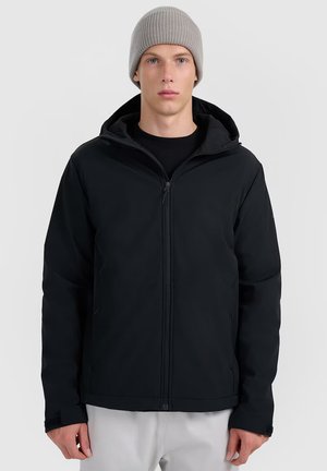 Young man wearing a grey knit beanie and a black zip-up hooded jacket, standing against a plain light background.