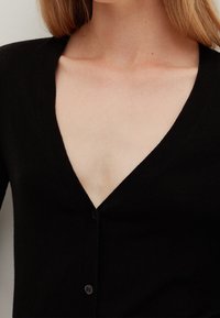 Černý pletený kardigan s hlubokým výstřihem do V a zapínáním na jeden knoflík na hrudi. Hladká textura, přiléhavý střih.