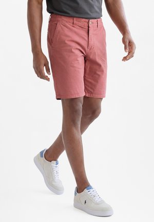 Mann trägt rosafarbene Shorts, ein dunkelgraues Shirt und weiße Sneakers und läuft auf einem einfarbigen weißen Hintergrund.