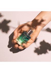 Grön och klar glasdroppeflaska med silverlock, märkt "ANNE SEMONIN," hållen i en hand mot en mjuk beige bakgrund.