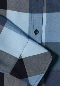 Gros plan d'une manche plissée de chemise à carreaux bleus et noirs avec une couture visible et un bouton noir sur le panneau avant.