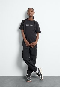 Svart t-shirt med "JORDAN" text, kombinerat med svarta cargobyxor som har fickor och elastiska manschetter. Skorna är svarta och vita sneakers.