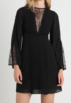 Vrouw die een zwarte jurk met lange mouwen draagt, met kanten panelen op de borst en mouwen, voorzien van geplooide details aan de voorkant.