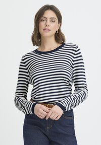 Chemise à manches longues rayée bleu marine et crème, avec un col rond et des poignets côtelés, associée à un jean foncé et une ceinture marron.