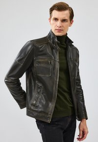 Veste en cuir noir avec un col haut, une poche zippée sur la poitrine et des poches latérales, et des coutures contrastées. Portée par-dessus un col roulé vert foncé.