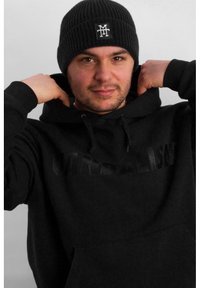 Black hoodie with "VANDALISK" in embossed text, styled with a ribbed black knit beanie featuring white logo, worn by a person pulling the hoodie.