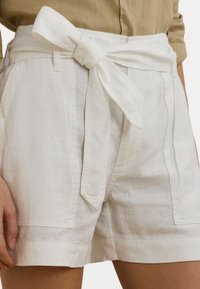 Baltos linines šortai su plačiu liemeniu, surištu dryžuotu mazgu ir šoninėmis kišenėmis, demonstruojantys laisvą stilių ir švelnią tekstūrą.