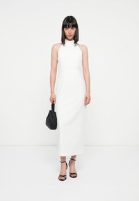 Witte halter-jurk met een aansluitende silhouet, een zachte textuur en midi-lengte. Gepaard met een zwarte handtas en zwarte sandalen met hakken en bandjes.