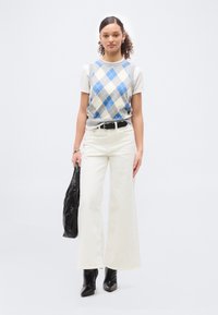 Γυναίκα που φοράει αμάνικο πουλόβερ σε μπλε και γκρι αργκόλ πάνω από λευκό T-shirt, λευκά παντελόνια φαρδιάς γραμμής, μαύρες μπότες, κρατώντας μαύρη τσάντα.