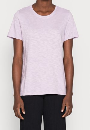 Person iført en lys lilla kortærmet t-shirt og sorte bukser, stående med armene afslappet ned langs siden mod en ensfarvet baggrund.