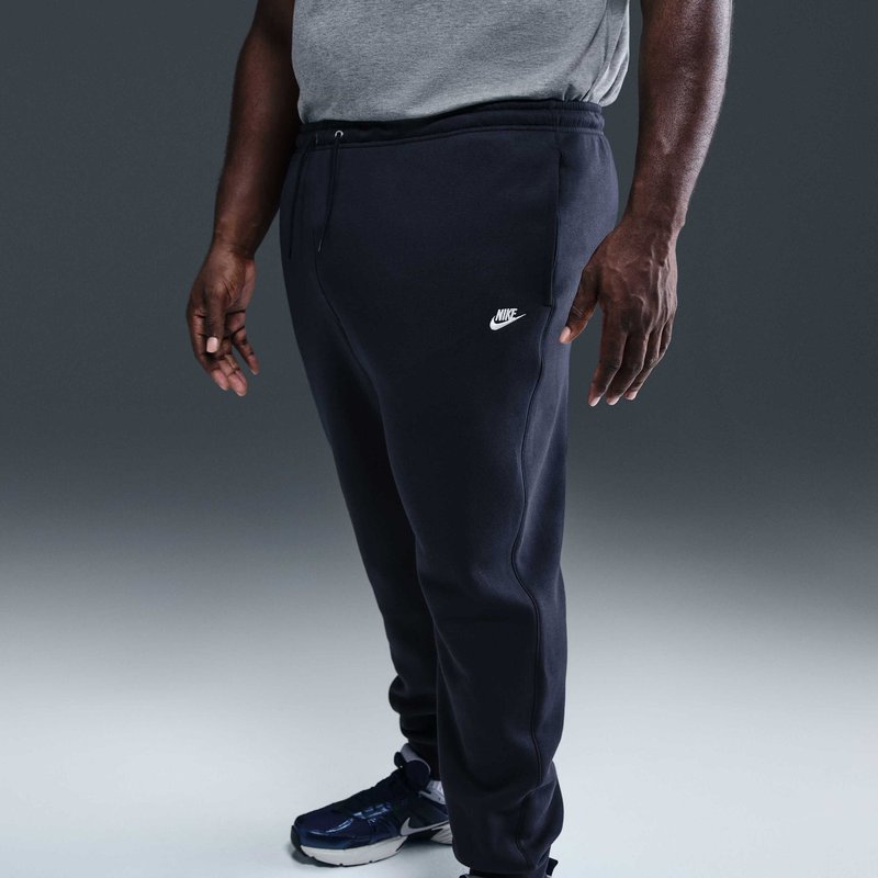 Uomo con pantaloni della tuta neri Nike, t-shirt grigia e scarpe da ginnastica nere in piedi contro uno sfondo grigio semplice, braccia rilassate lungo i fianchi.