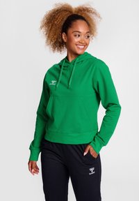 Grüne Kapuzenjacke mit Vordertasche und Kordelzug. Aus weichem Stoff gefertigt; mit einem Logo auf der linken Brust. Kombiniert mit schwarzen Hosen.