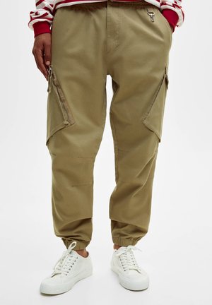 Persona che indossa pantaloni cargo color cachi con polsini elasticizzati, tasche laterali con cerniera e scarpe da ginnastica bianche con lacci. Mani parzialmente nelle tasche.