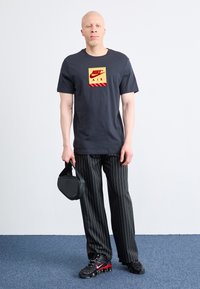 Μαύρη μπλούζα με το λογότυπο «Nike Air» σε κόκκινες και χρυσές αποχρώσεις, συνδυασμένη με μαύρο παντελόνι με ρίγες και μαύρες παπούτσια με κόκκινες λεπτομέρειες, κρατώντας μία μαύρη τσάντα.