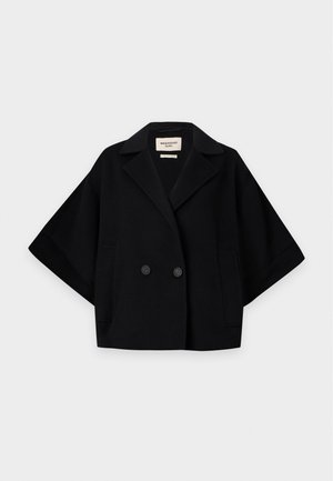 Kvinde med kort mørkt hår iført sort oversized jakke, hvid langærmet top, sorte bukser og beige høje hæle, står foran hvid baggrund.