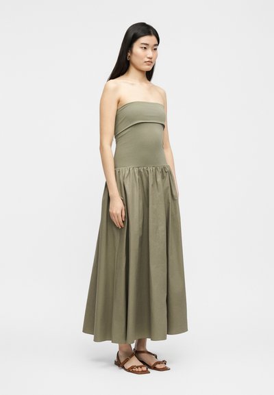 Γυναίκα με μακριά σκούρα μαλλιά, ντυμένη με μάξι φόρεμα ανοιχτού ελιάς χωρίς τιράντες και καφέ επίπεδα σανδάλια, στέκεται μπροστά σε απλό φωτεινό φόντο.