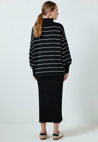 Fekete-fehér csíkos, oversized pulóver magas nyakkal, fekete bordázott midi szoknyával és világos színű cipőkkel párosítva.