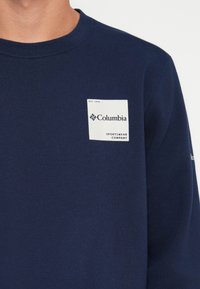Felpa navy con scollo rotondo, dotata di una patch in tessuto bianco che riporta la scritta "Columbia Sportswear Company" e "EST. 1938" in testo nero.