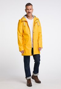 Cămașă de raincoat impermeabilă galbenă, cu glugă cu șnur, două buzunare frontale și accesorii metalice argintii, purtată cu blugi negri și cizme maro.