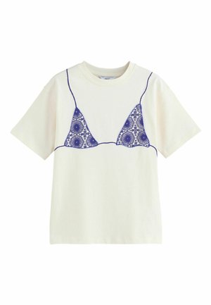 Ung kvinde med mørkt, bølget hår bærer en hvid t-shirt med et lilla blonder-bh design og grønstribede shorts, stående mod en stenvæg.