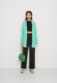 Cardigan vert menthe à l'avant ouvert, à manches longues, porté sur un haut noir court et un pantalon noir large. Les accessoires comprennent un sac vert.