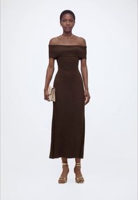 Γυναίκα με καφέ midi φόρεμα χωρίς ώμους, κρατώντας ένα μπεζ τσαντάκι και χρυσά ψηλοτάκουνα σανδάλια μπροστά σε απλό φόντο.