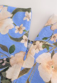 Tissu bleu avec imprimé floral beige et rose clair, comportant des feuilles vertes et une fermeture éclair bleue partiellement ouverte au centre.