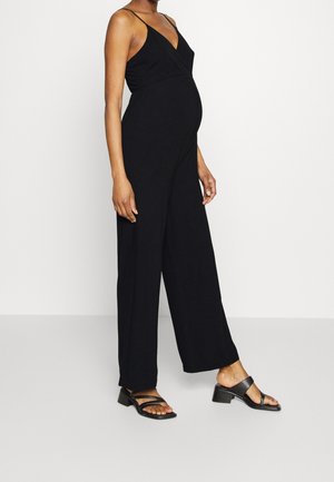 Czarny kombinezon z dekoltem w serek, regulowanymi cienkimi ramiączkami i szerokimi nogawkami. Materiał jest lekki i gładki, w połączeniu z czarnymi sandałami na paski.