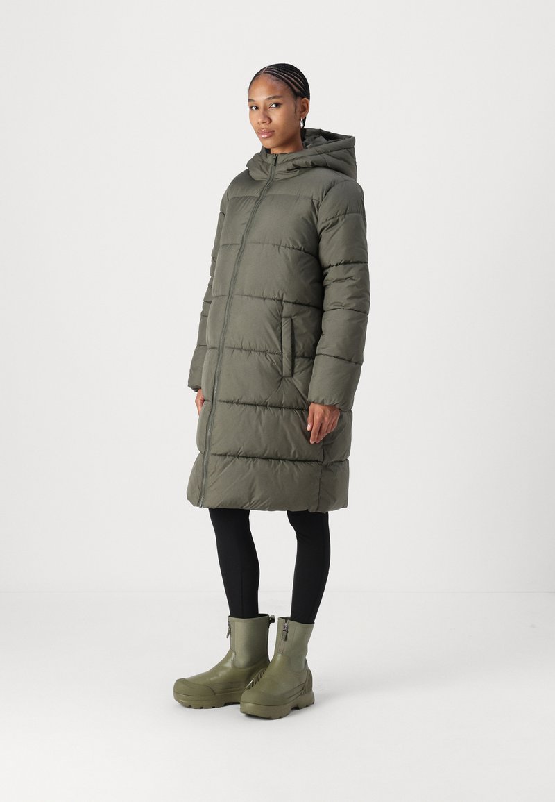 Cappotto lungo imbottito verde oliva con cappuccio, cuciture verticali, zipper frontale e tasche laterali; abbinato a stivali verdi alla caviglia e leggings neri.