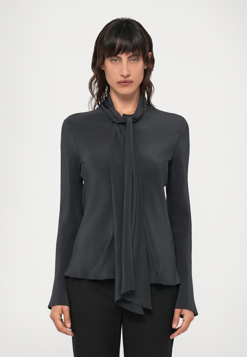 Dunkelgraue Bluse mit einem Bindungsdetail am Hals, langen Ärmeln und einem glatten, fließenden Stoff, der sanft über den Körper fällt.