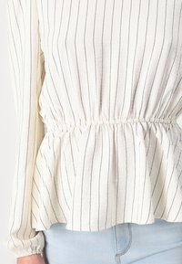 Top blanc avec des rayures noires verticales, présentant un design à peplum et une taille froncée. Manches longues avec poignets élastiques, en tissu léger.