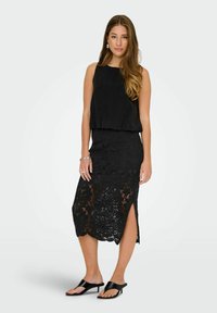 Schwarzes, ärmelloses Oberteil mit lockerer Passform, kombiniert mit einem schwarzen Spitzen-Midikleid mit floralen Mustern und einem seitlichen Schlitz. Getragen mit schwarzen Sandalen.