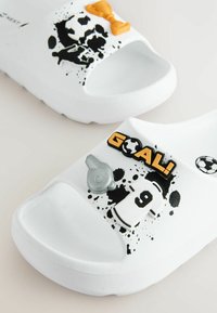 Des sandales blanches à glissière ornées de décorations sur le thème du football en noir et orange, comprenant un trophée, un ballon de football, un sifflet et un maillot avec le numéro 9.