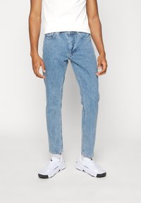 Ljusblå denimjeans, tapered fit, slitningar vid knäna, matchat med vita sneakers. Slät textur, klassisk femfickorsdesign.