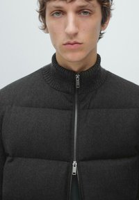 Young person with light eyes wearing a dark padded jacket with a high ribbed collar and a partially zipped front.