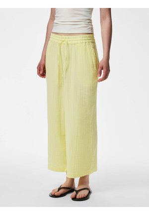 Femme portant un pantalon jaune ample à cordon, un haut court blanc et des tongs noires, debout devant un fond uni.